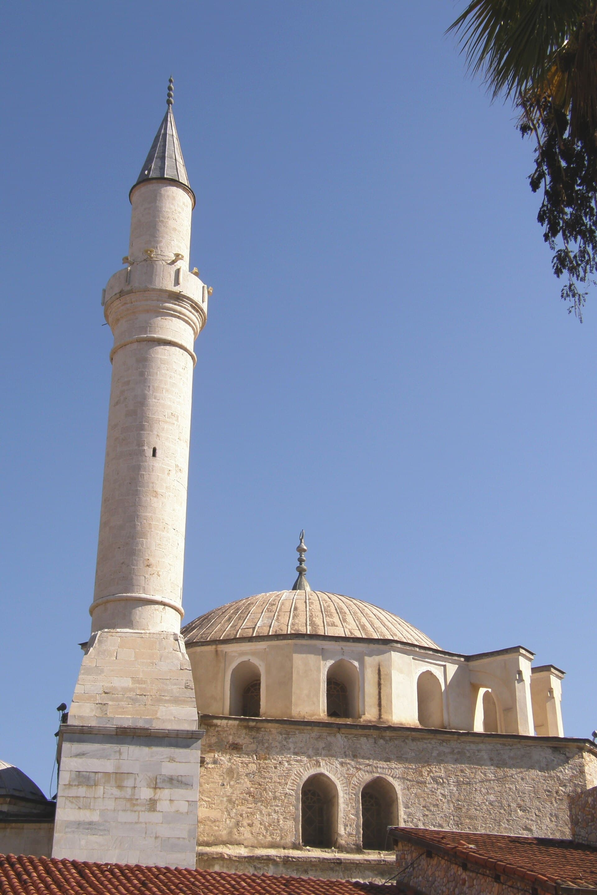 Looking up at the stone minaret and ribbed lead dome of the Ottoman-era Kaleiçi Mosque (Öküz Mehmet Paşa Camii) in Kuşadası's old town, set against a clear blue sky with palm fronds at the frame's edge.