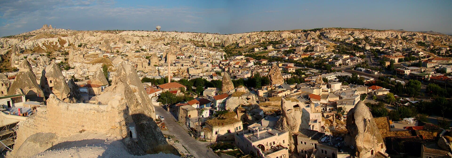 Panoramic view over Göreme, Cappadocia, with houses built among fairy-chimney rock formations and a hot air balloon near the ridge in the distance