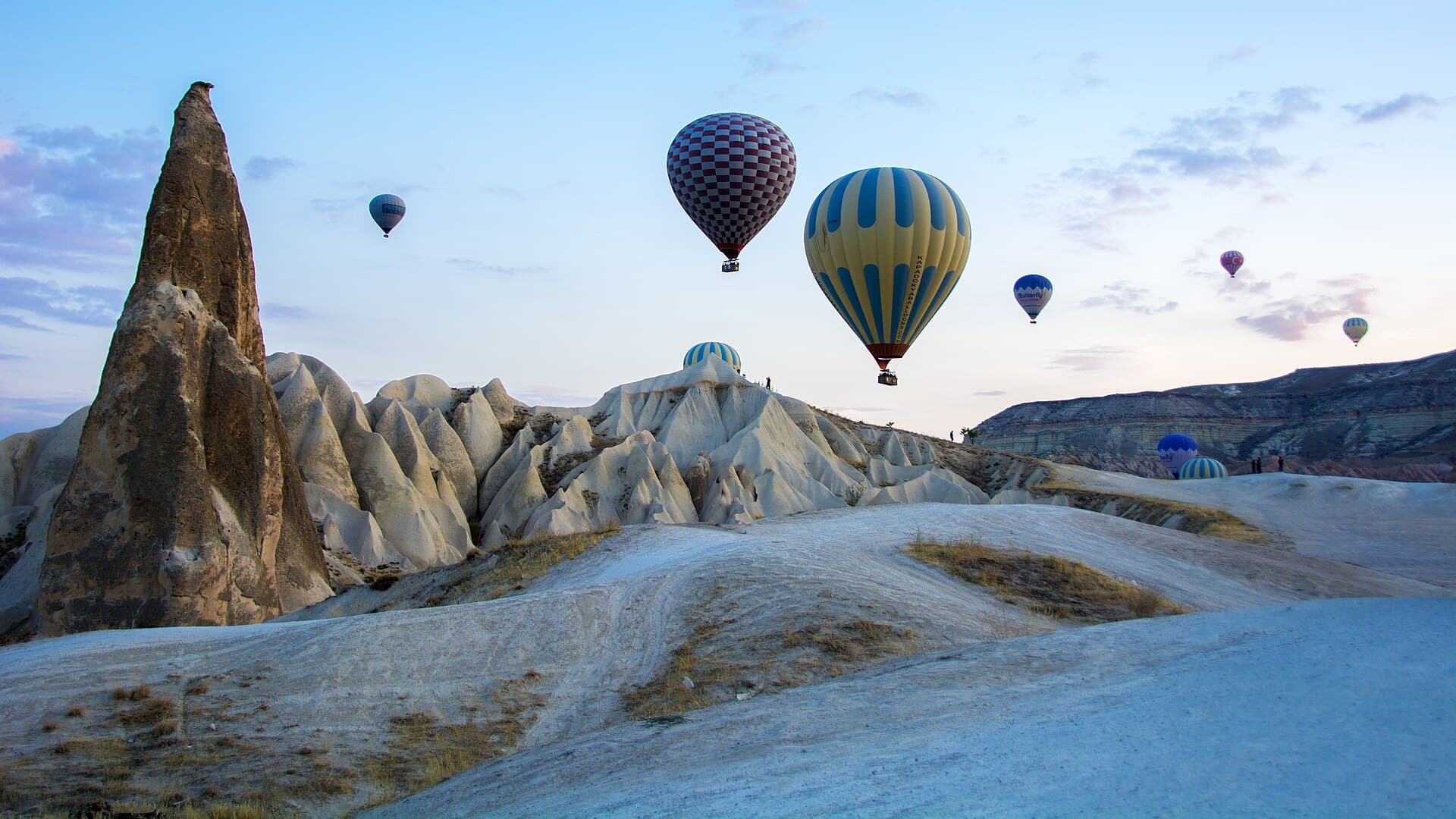 Hot air balloons rise beside a tall fairy chimney rock spire as more balloons drift across the dawn sky over Cappadocia