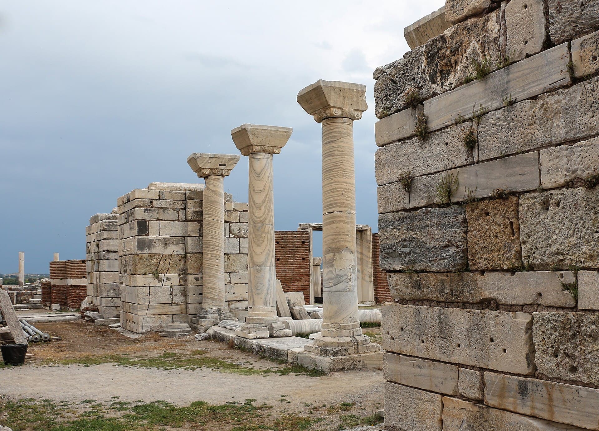 Three tall re-erected marble columns with carved Ionic-style capitals stand among the ruined stone and brick walls of the Basilica of St. John in Ephesus, under a partly cloudy sky.