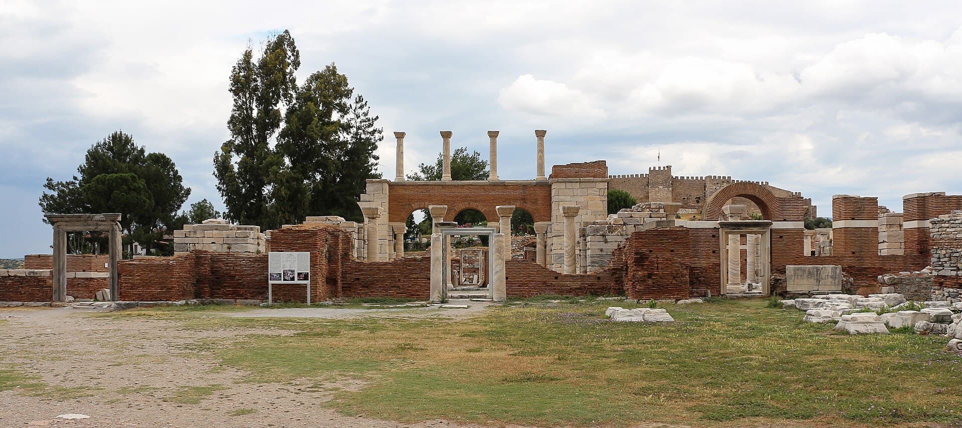 A wide view across the grassy ruined footprint of the Basilica of St. John on Ayasuluk Hill, showing brick walls, an arched colonnade with re-erected columns, and the battlements of Ayasuluk (Selçuk) Castle visible on the hilltop behind.