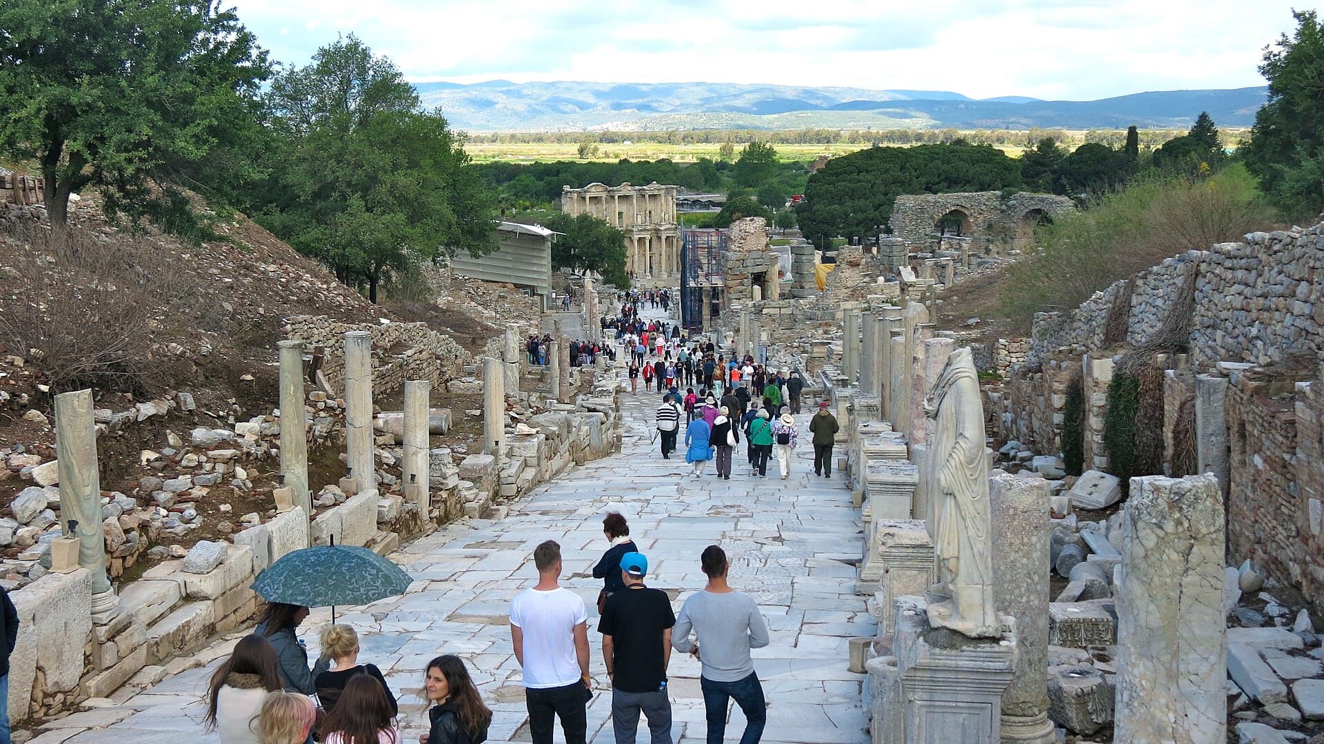 A downhill view along the marble-paved Curetes Street at Ephesus, lined with columns and ruined building facades on both sides, with the two-story facade of the Library of Celsus visible in the distance and tourists walking down the slope.