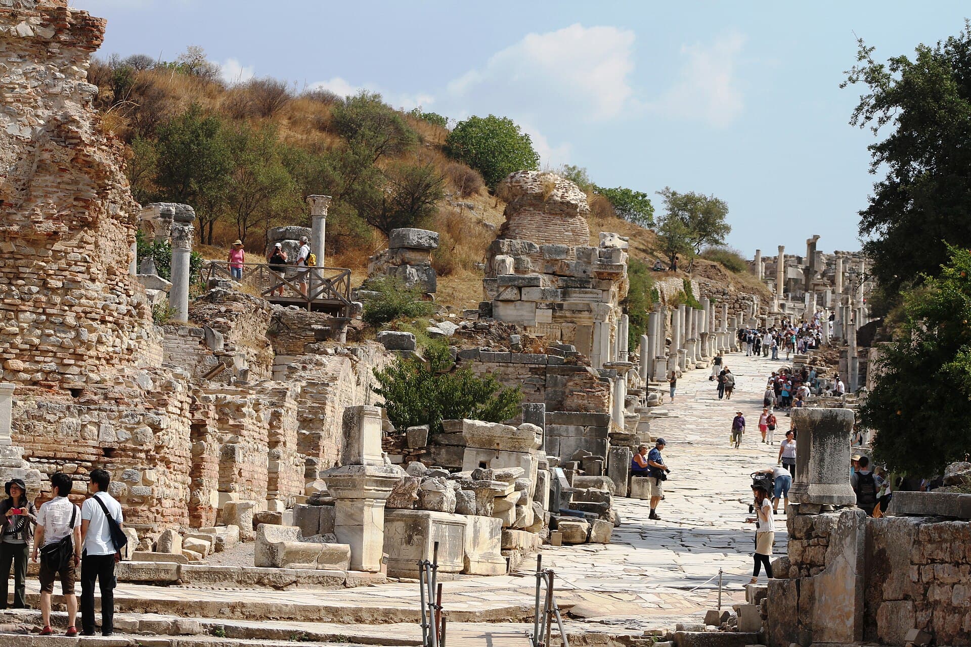 An angled view up Curetes Street at Ephesus, with marble columns lining the paved street on the right, the ruined brick and stone walls of the Terrace Houses on the left, and tourists walking among the ruins.