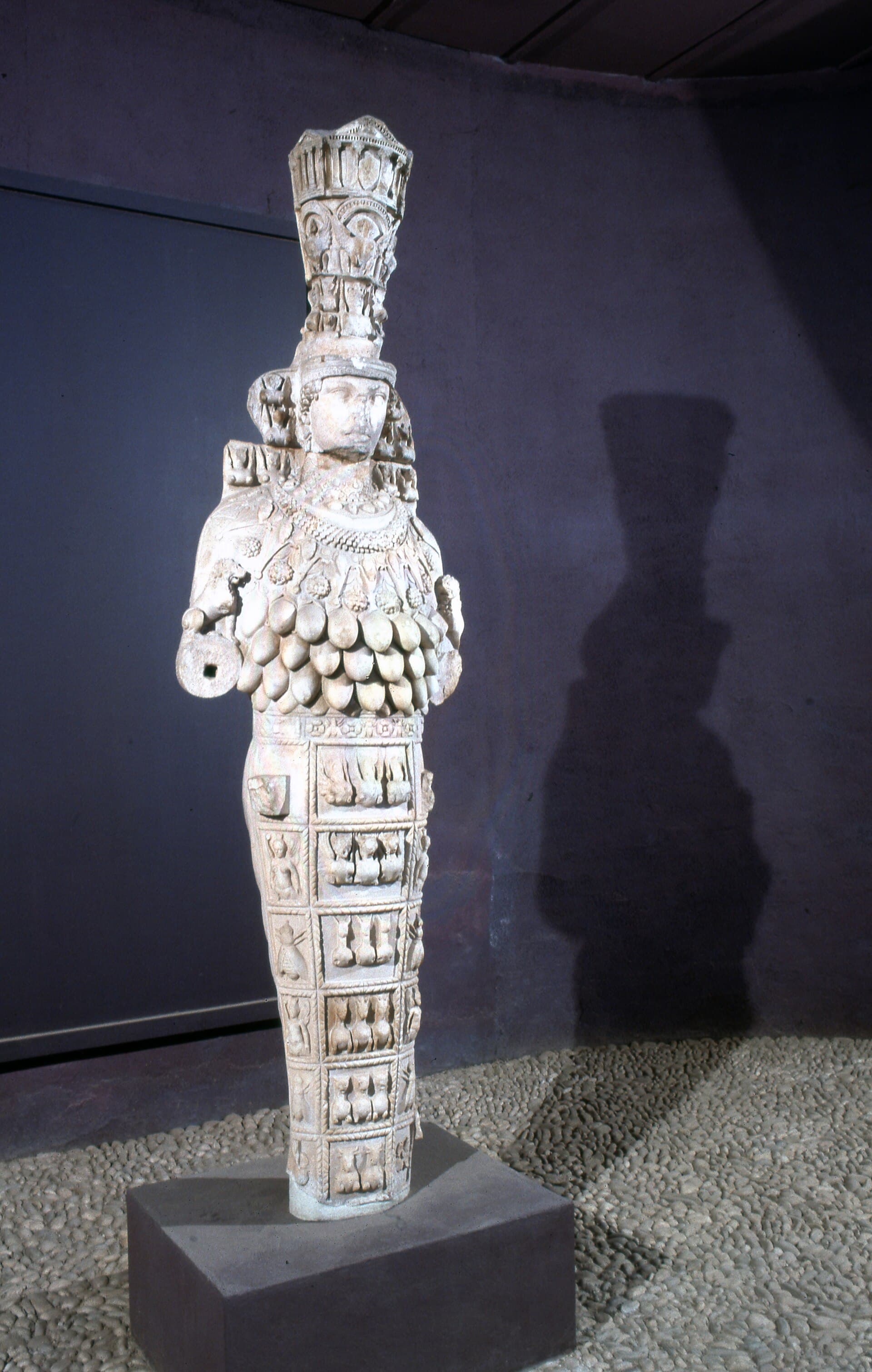The "Great Artemis" (Colossal Artemis) statue at the Ephesus Archaeological Museum in Selçuk, Turkey — a weathered marble cult statue shown full-length with a tall tiered headdress, an ornate collar, rows of round egg-like ornaments down the torso, and a paneled skirt, standing in a dark museum gallery.