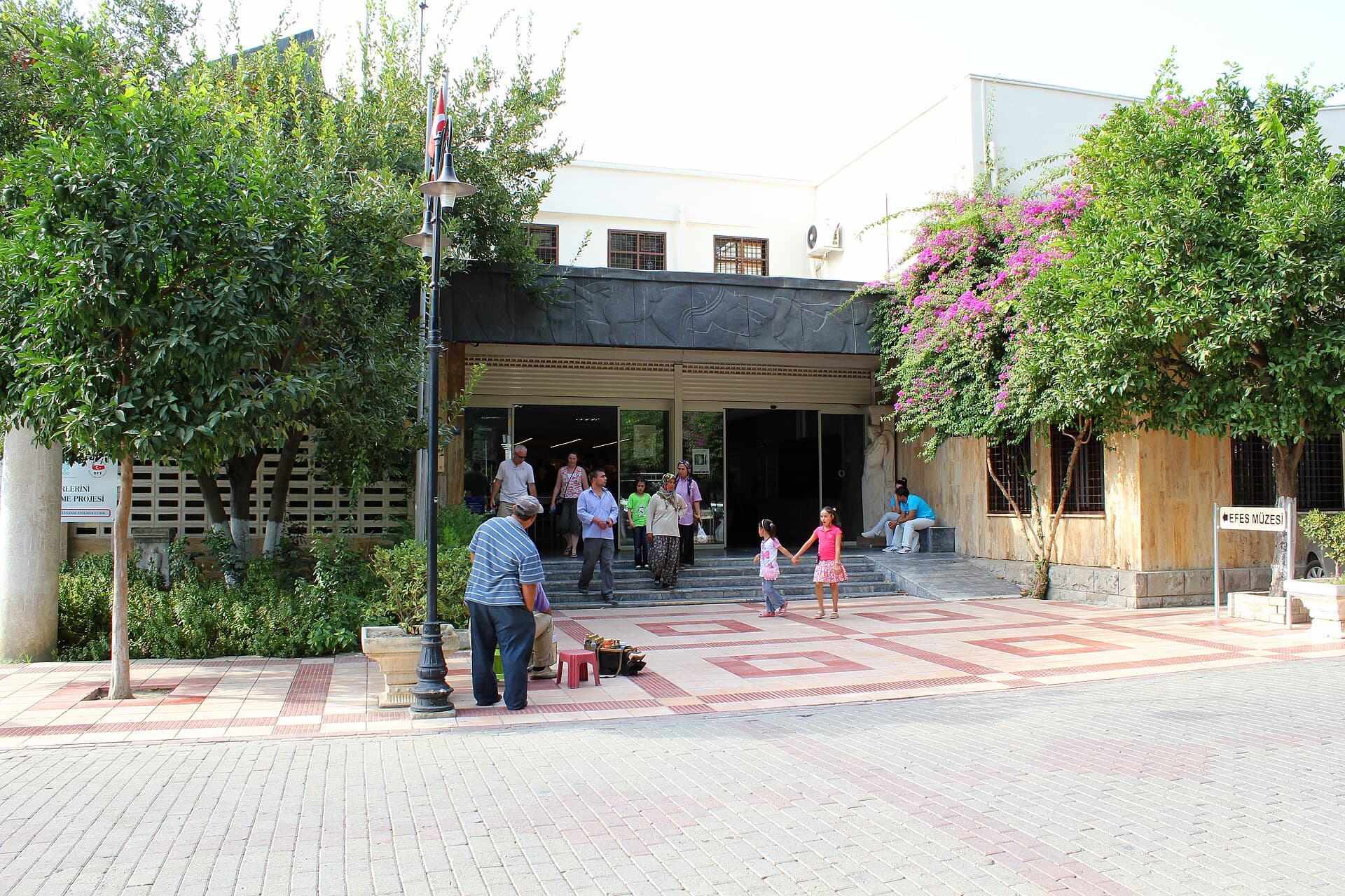The entrance to the Ephesus Archaeological Museum in Selçuk, Turkey — a low modern building with a dark stone relief panel above the doorway, visitors walking through the entrance, and trees and pink bougainvillea flanking the courtyard.