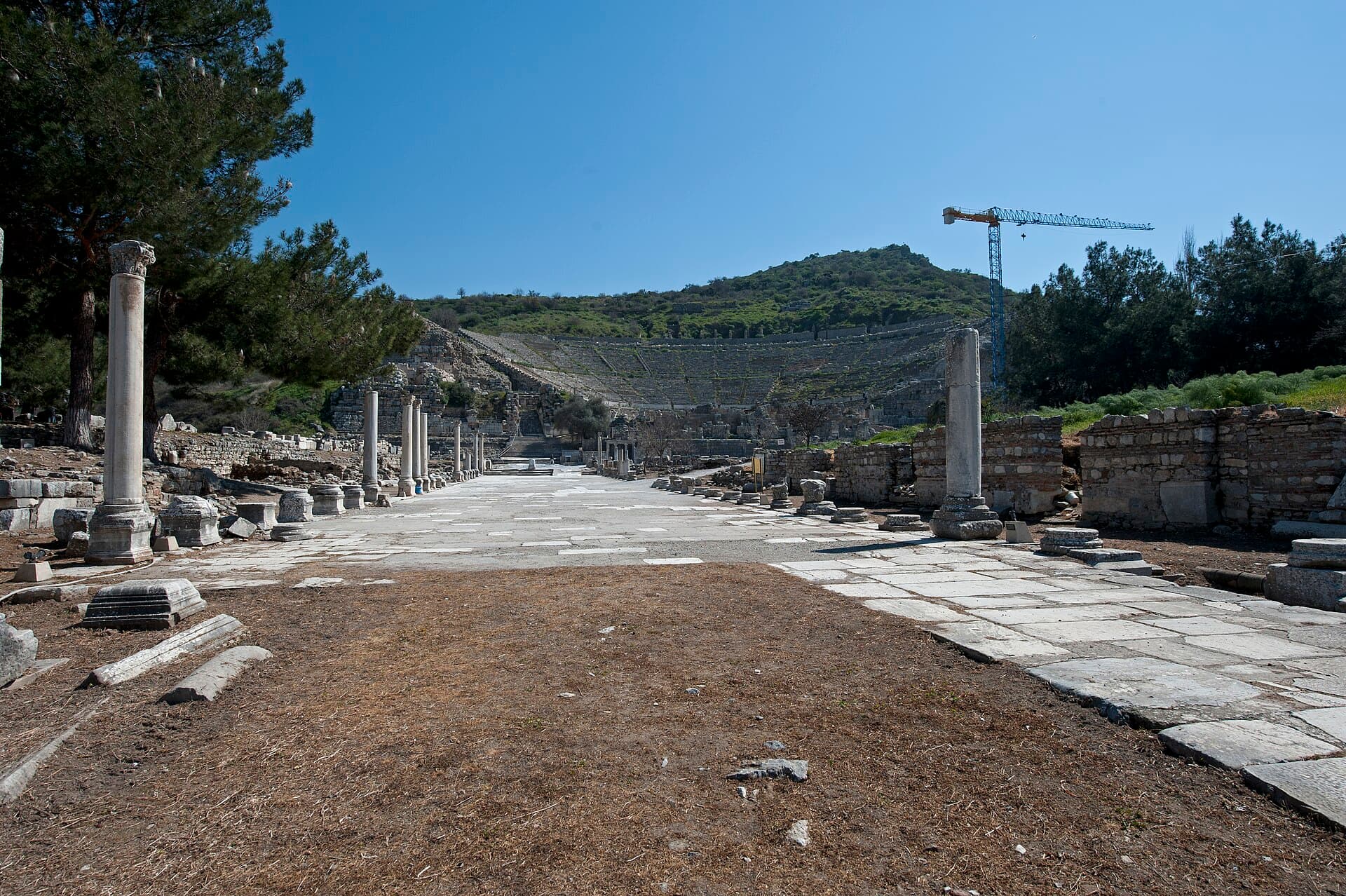 Colonnaded Arcadiane (Harbour) Street at Ephesus leading toward the Great Theatre, whose tiered stone seating rises into the green hillside in the distance.