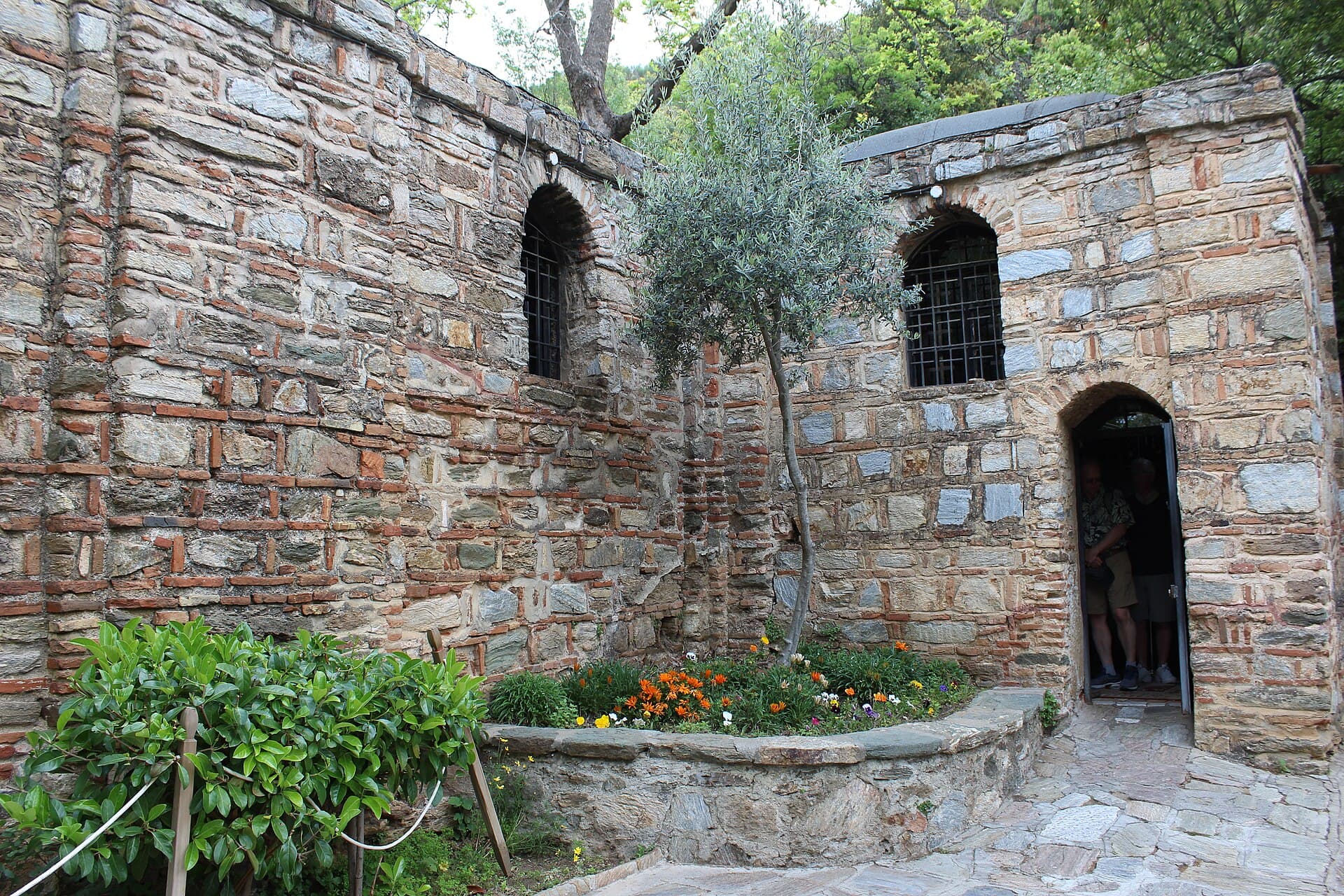 Exterior stone wall of the House of the Virgin Mary chapel near Ephesus, Turkey, showing two barred windows, an open doorway with visitors inside, and an olive tree growing beside the entrance.