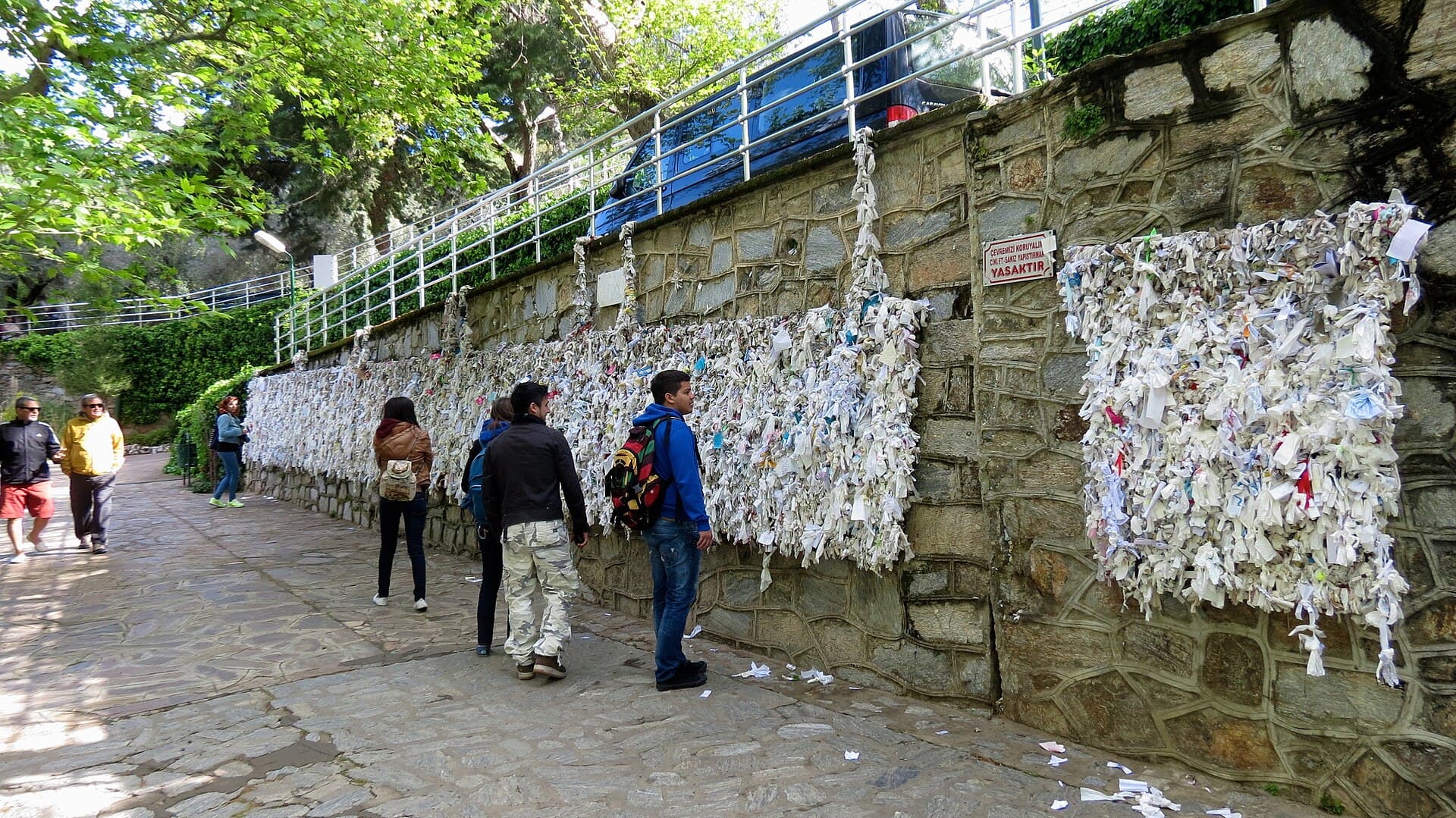 The Wishing Wall below the House of the Virgin Mary near Ephesus, a long stone wall densely covered with white paper and cloth prayer notes left by visitors, who walk along the path beside it.