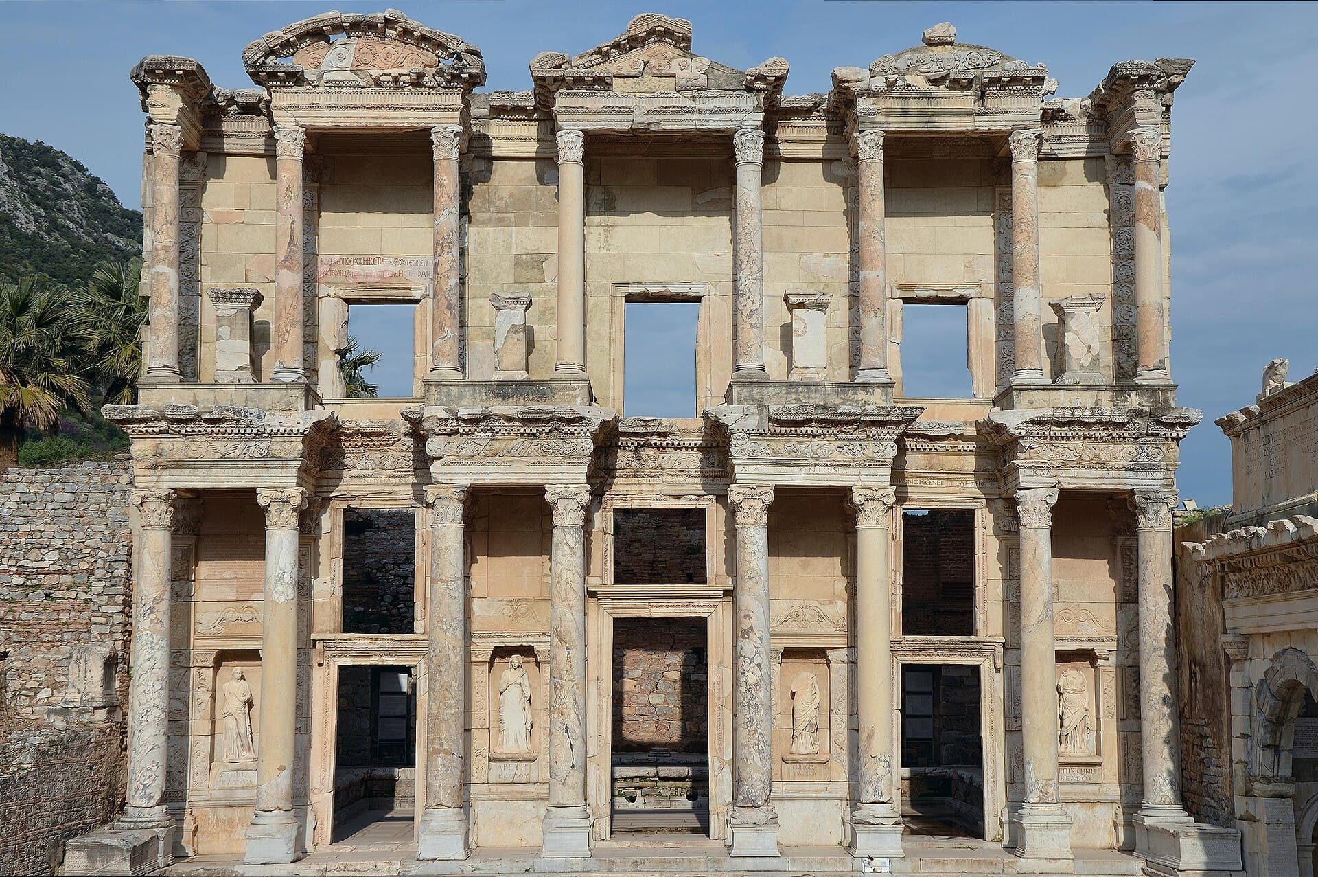 Straight-on view of the fully reconstructed two-storey marble facade of the Library of Celsus at Ephesus, with all four Corinthian-columned bays, the four statue niches, and a clear blue sky behind, wooded hillside to the left.
