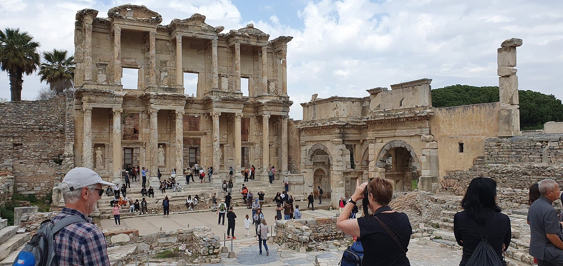 Wide angle view of the Library of Celsus facade beside the ruined Gate of Mazeus and Mithridates at Ephesus, with tourists gathered on the plaza and stairs below and palm trees at the edge of the frame.