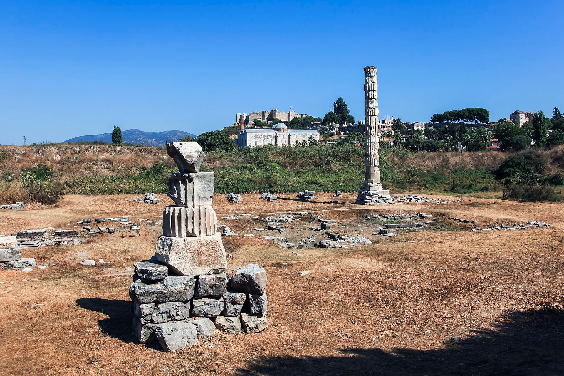 A wide daytime view of the sparse, grassy Temple of Artemis archaeological site near Selçuk, Turkey, showing a single tall reassembled column and a smaller pile of column fragments in an open field, with the Isa Bey Mosque and the hilltop Ayasuluk Citadel visible in the background.