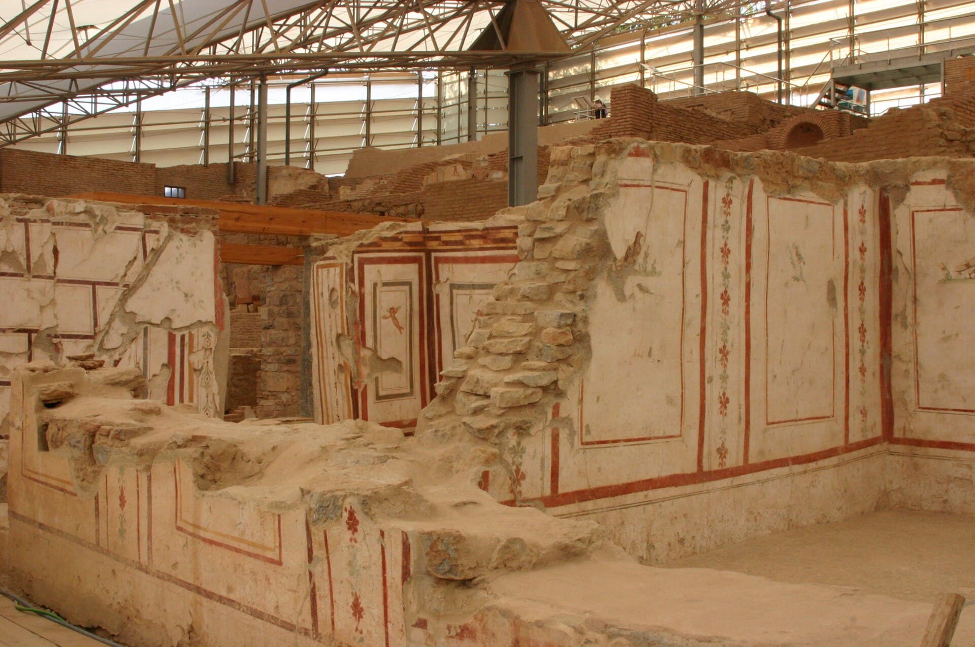 Close-up of Roman-era frescoed walls in the Terrace Houses at Ephesus, showing red-bordered decorative panels with floral garland motifs, preserved beneath the site’s protective roof structure.