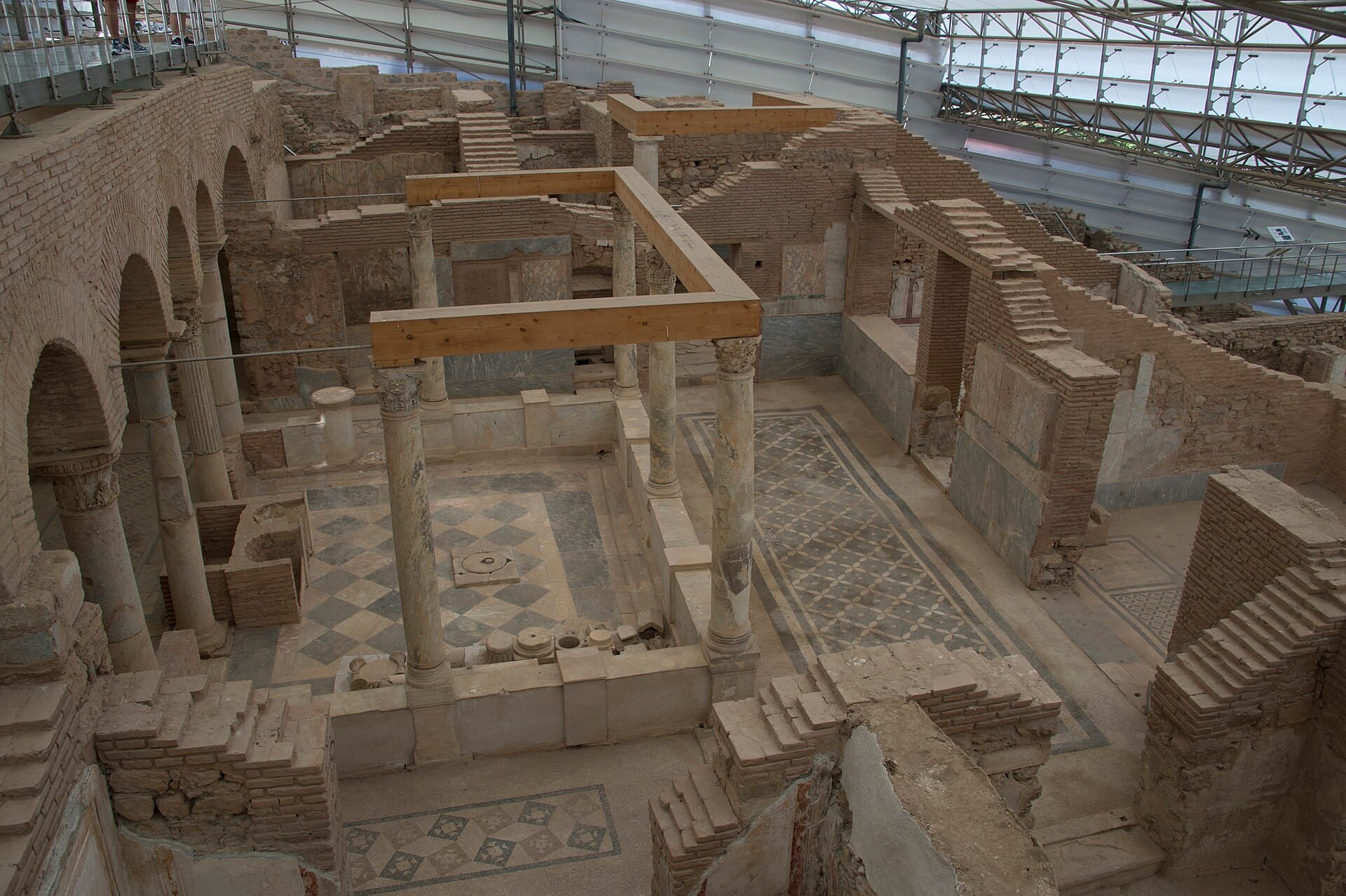 Elevated interior view of the Terrace Houses at Ephesus, showing excavated Roman rooms with geometric black-and-white mosaic floors and marble columns, with visitors on the raised walkway beneath the site’s glass-and-steel roof canopy.