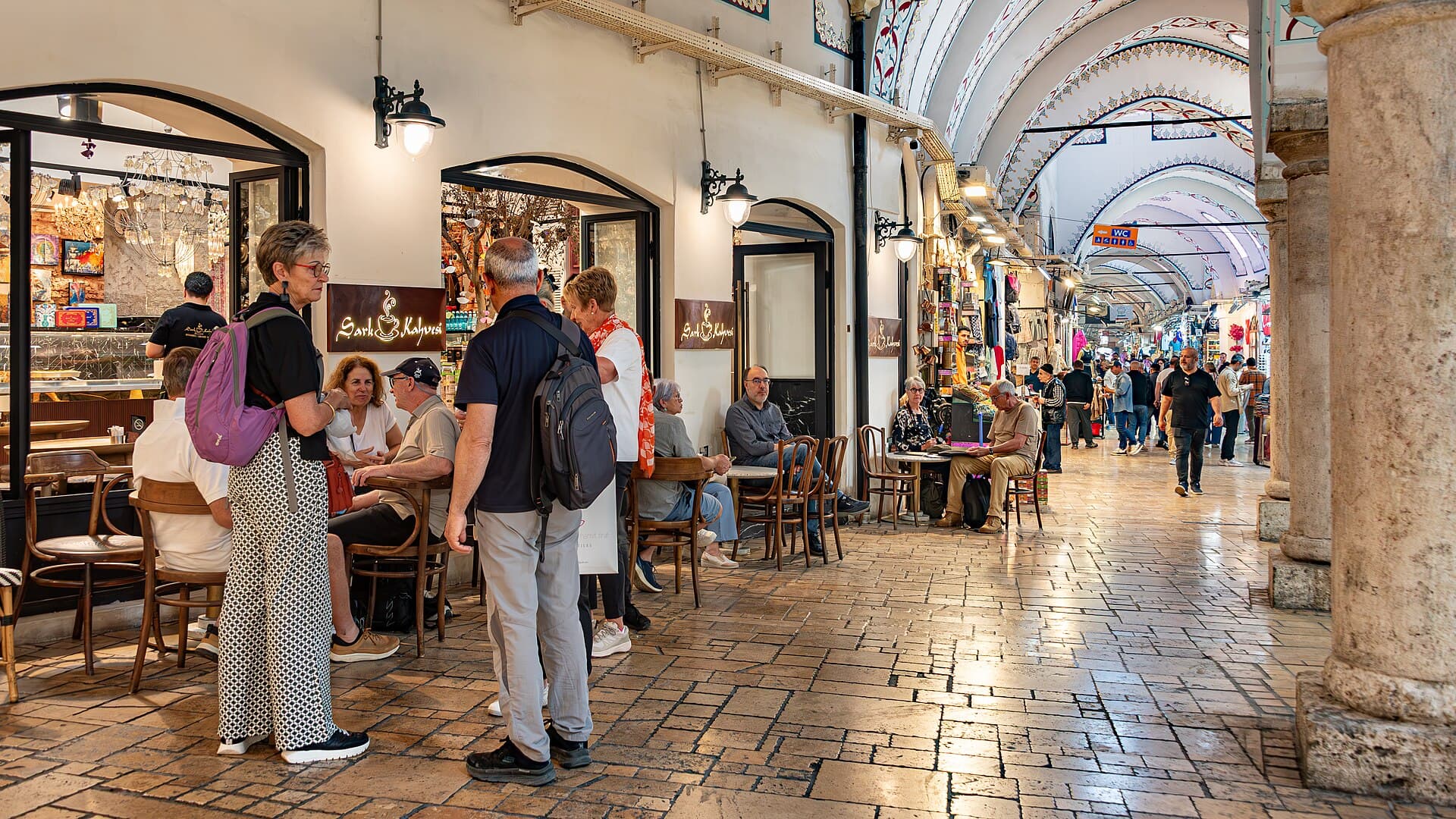 Shoppers and cafe tables beneath a decorated archway in a side aisle of the Grand Bazaar, with the main covered corridor visible beyond
