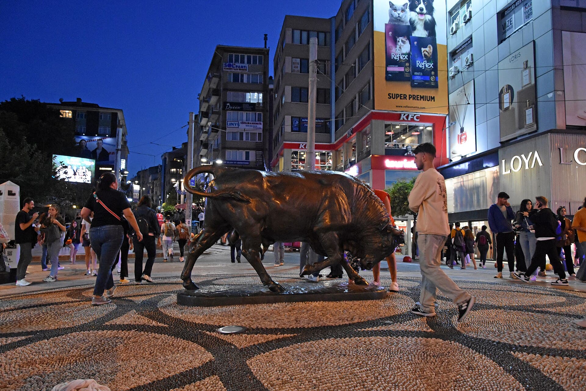Bronze bull statue (Boğa Heykeli) at the Altıyol intersection in Kadıköy, illuminated at dusk with pedestrians crossing the square