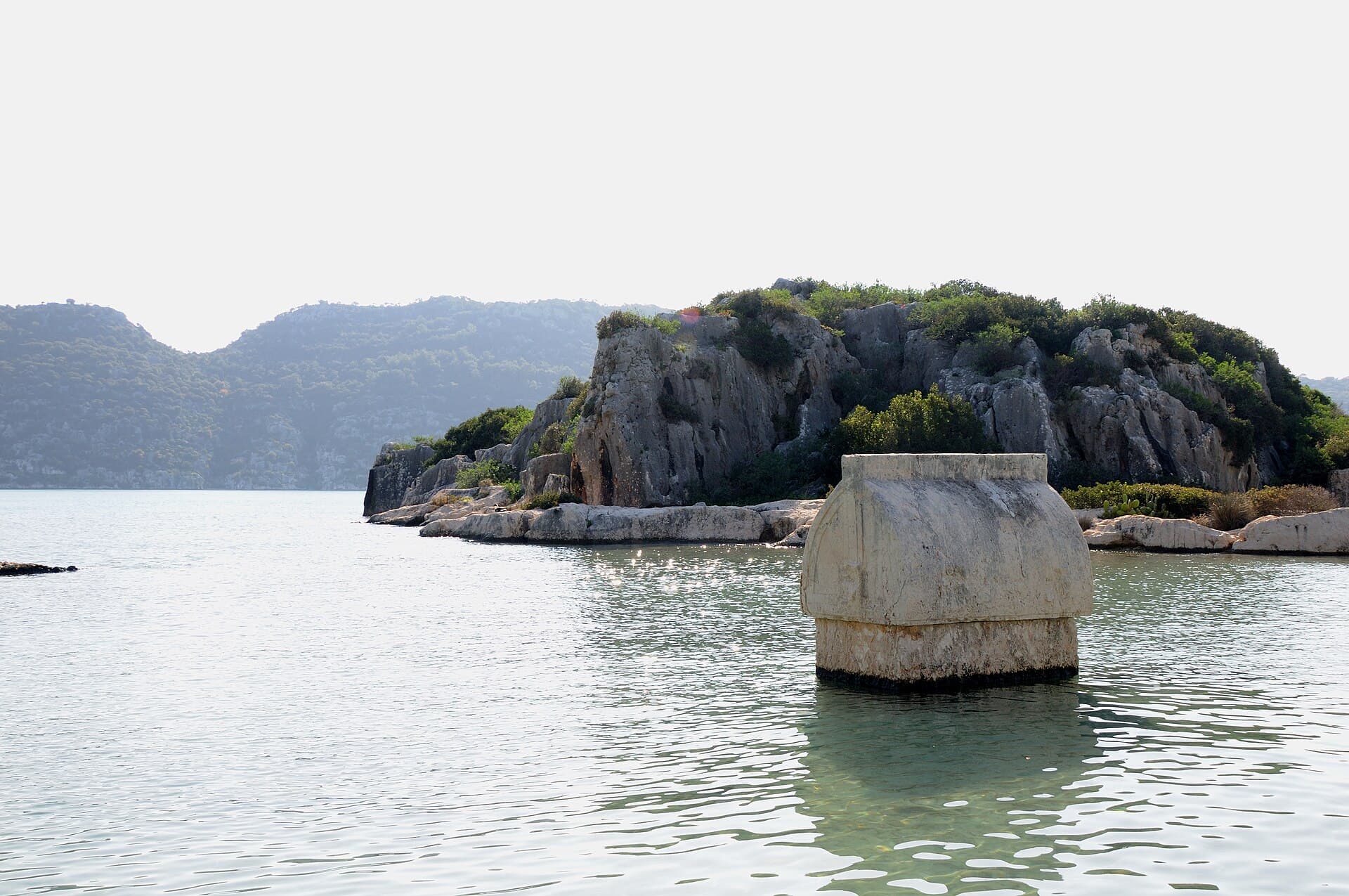 Lycian stone sarcophagus half-submerged in clear water off Kaleköy's rocky shore.