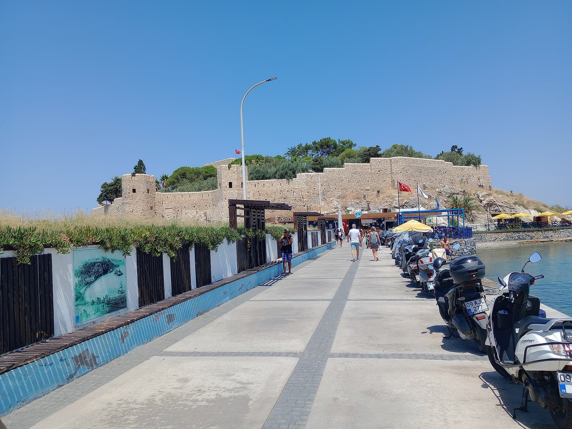 Visitors walking along the stone causeway to Güvercinada Kalesi in Kuşadası, with the fortress's stone walls, a Turkish flag, and scooters parked along the seaside railing under a clear blue sky.