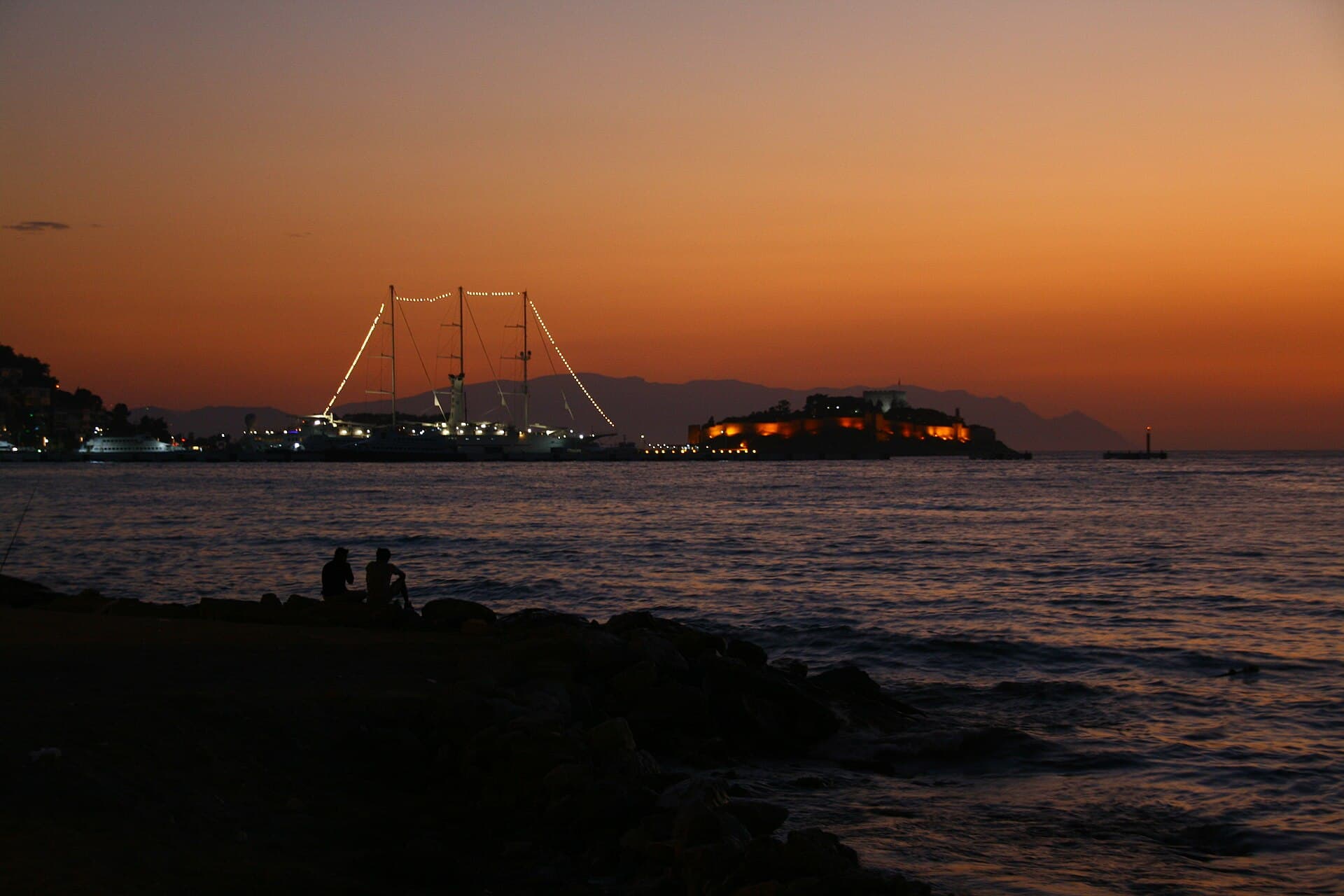 Dusk view across the water in Kuşadası showing Güvercinada's fortress walls lit in warm orange light, a light-strung tour boat near the town shoreline, and two people silhouetted on the rocks in the foreground.