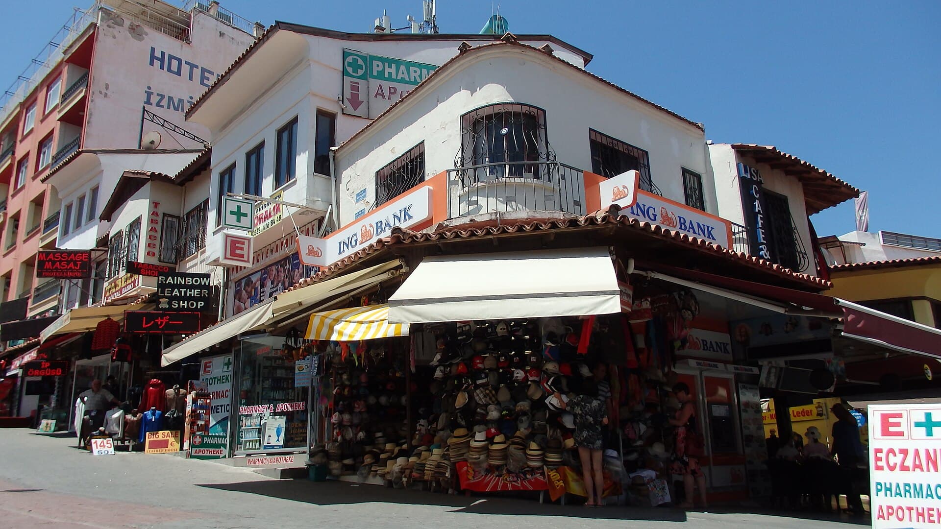A sunlit corner of Kuşadası's old-town bazaar streets, with a hat shop, leather-goods store, and pharmacy and bank signage under striped awnings, and tourists browsing on the paved corner.