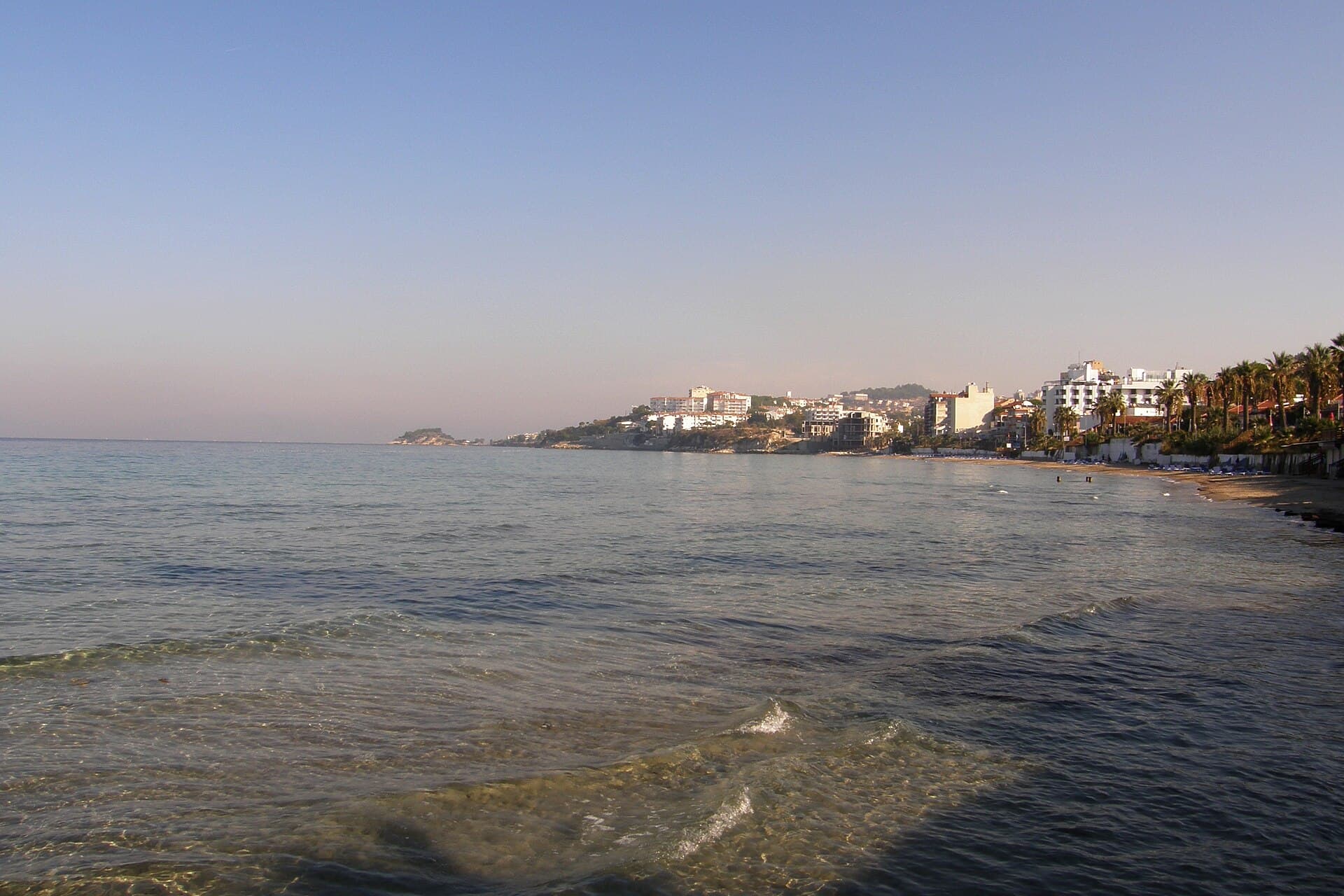 Daytime view across the calm sea of Ladies Beach (Kadınlar Denizi) in Kuşadası toward the curving shoreline, backed by white hotels and palm trees on the slope above the beach, with a rocky headland visible in the distance.