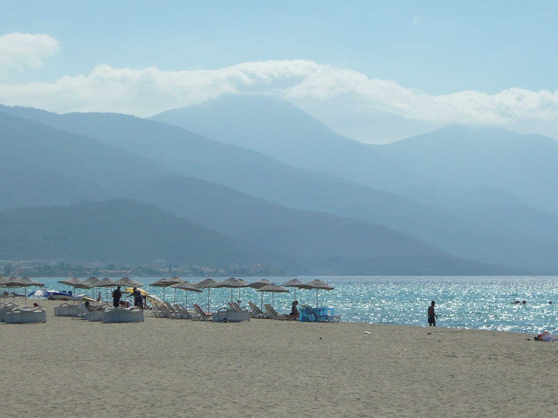 Straw umbrellas and white sun loungers line a wide sandy beach on Kuşadası's Long Beach near Davutlar, with calm, shallow turquoise water glittering in sunlight and hazy mountains rising across the gulf.