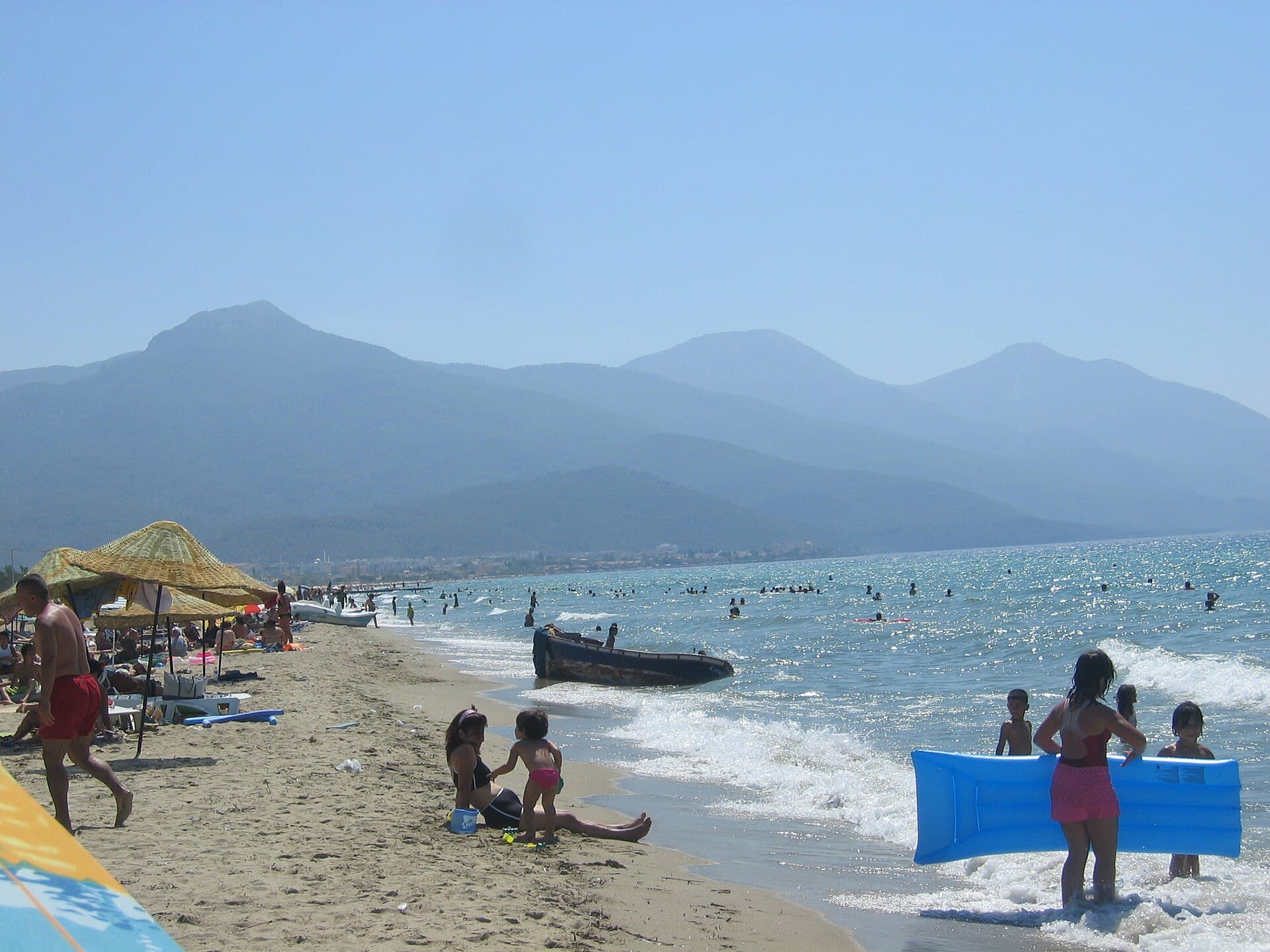 Bathers wade far out into shallow water off a curving sandy beach along Kuşadası's Long Beach near Davutlar, with a small wooden boat beached on the sand, a straw umbrella, and hazy mountains across the water.