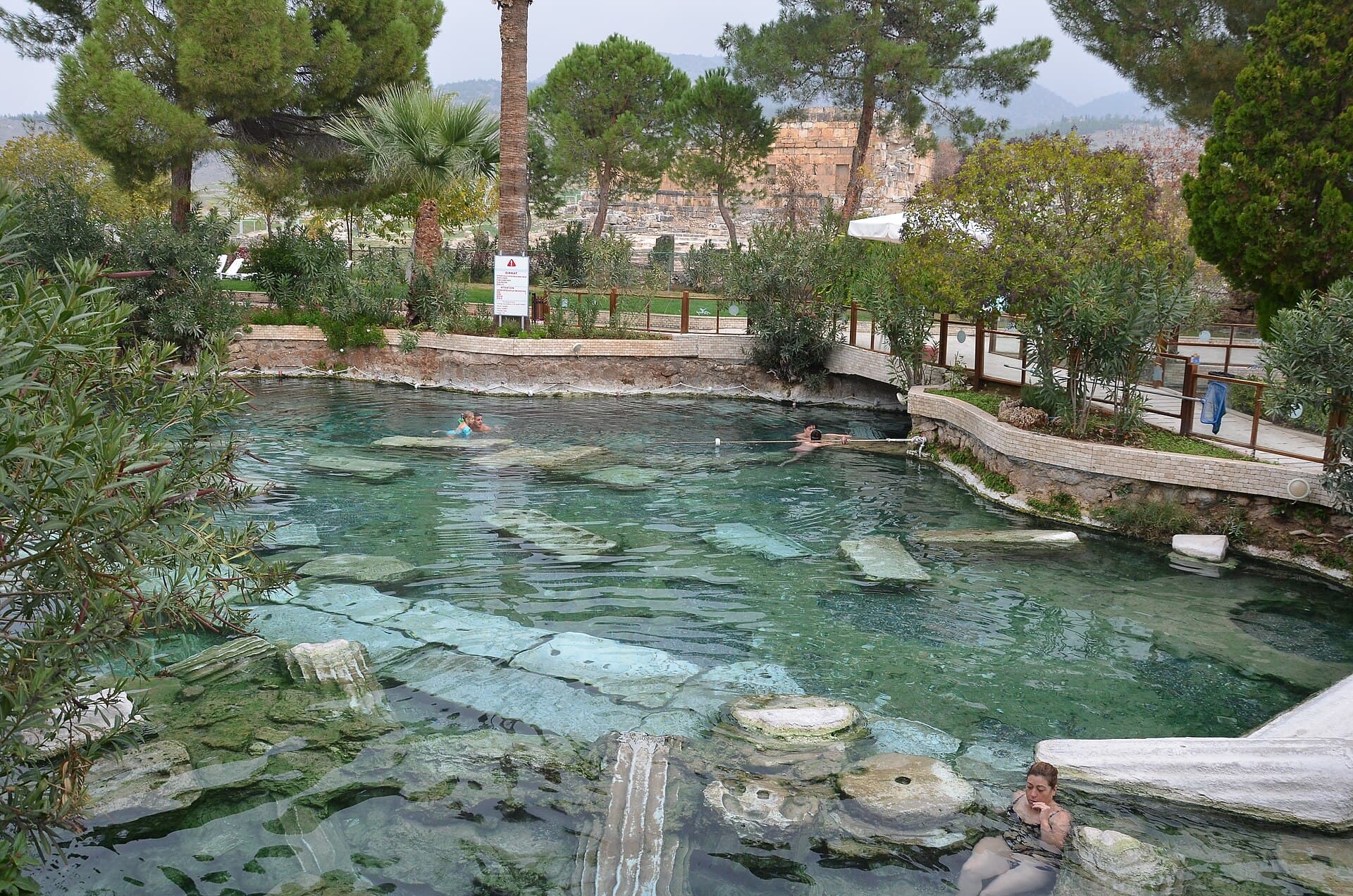 The Antique Pool at Hierapolis with fallen marble column drums and architrave blocks lying on the bottom of its clear turquoise thermal water, a few bathers swimming among them and pine-shaded gardens with ancient ruins beyond.