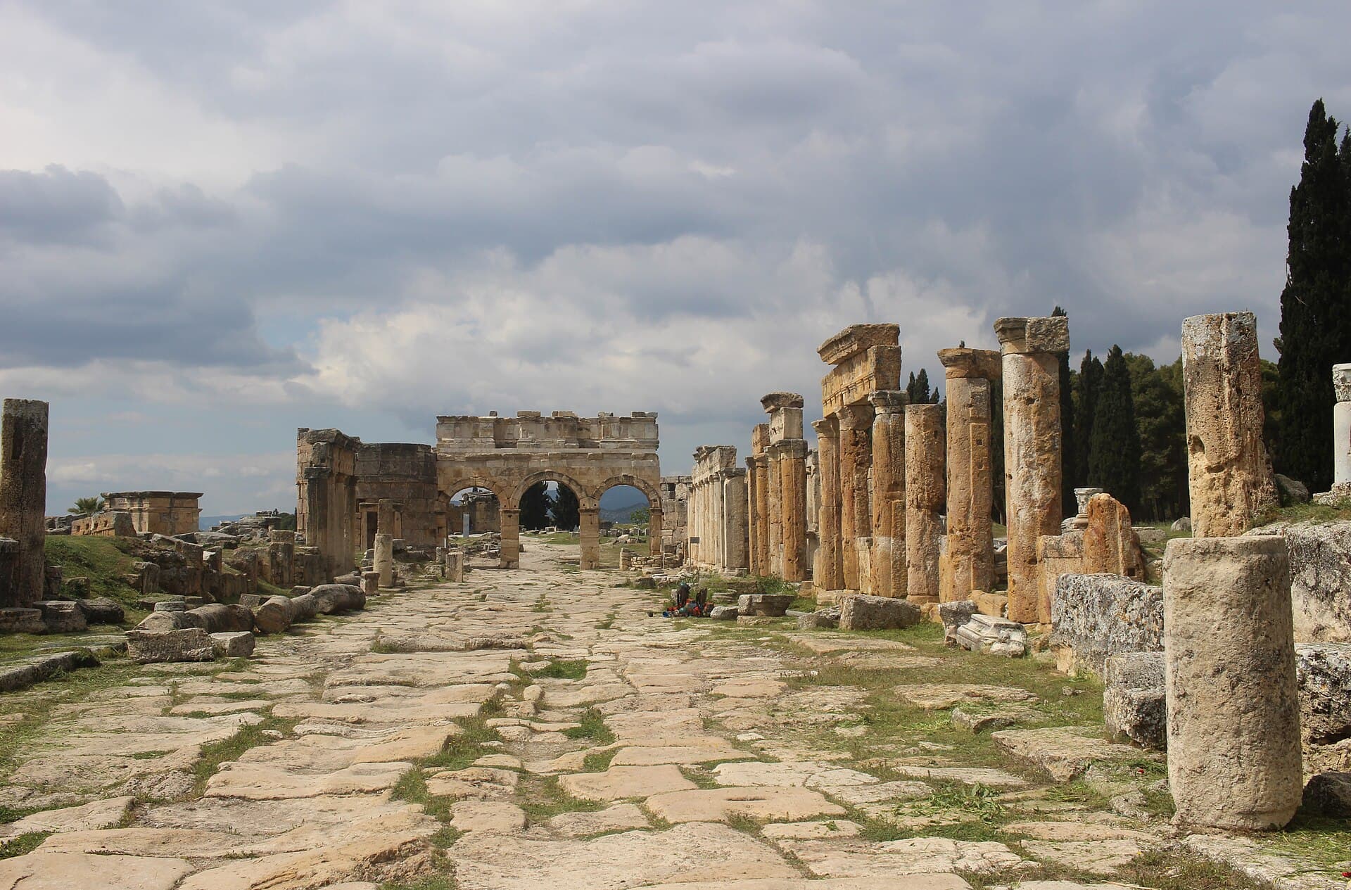 The wide stone-paved Frontinus Street at Hierapolis, lined with a colonnade of weathered columns on one side and closed at its far end by the triple-arched Frontinus Gate under a cloudy sky.