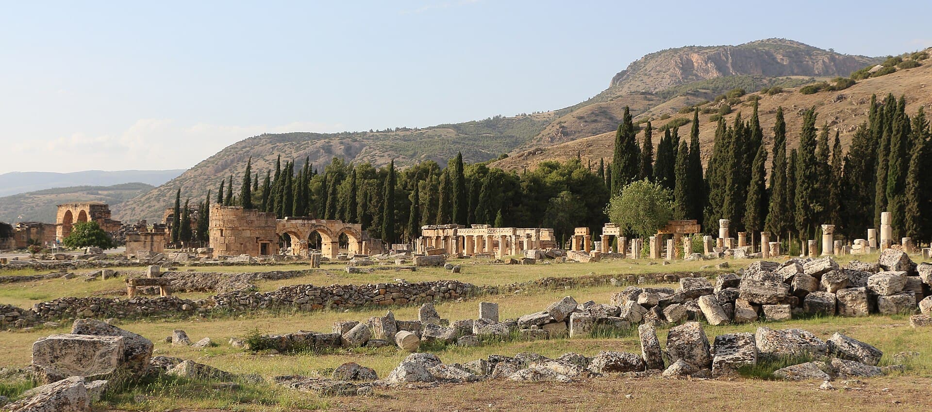 Wide evening view across the grassy plateau of Hierapolis, strewn with fallen stone blocks, with the arched gate ruins and colonnades of the ancient city among dark cypress trees below dry hills.