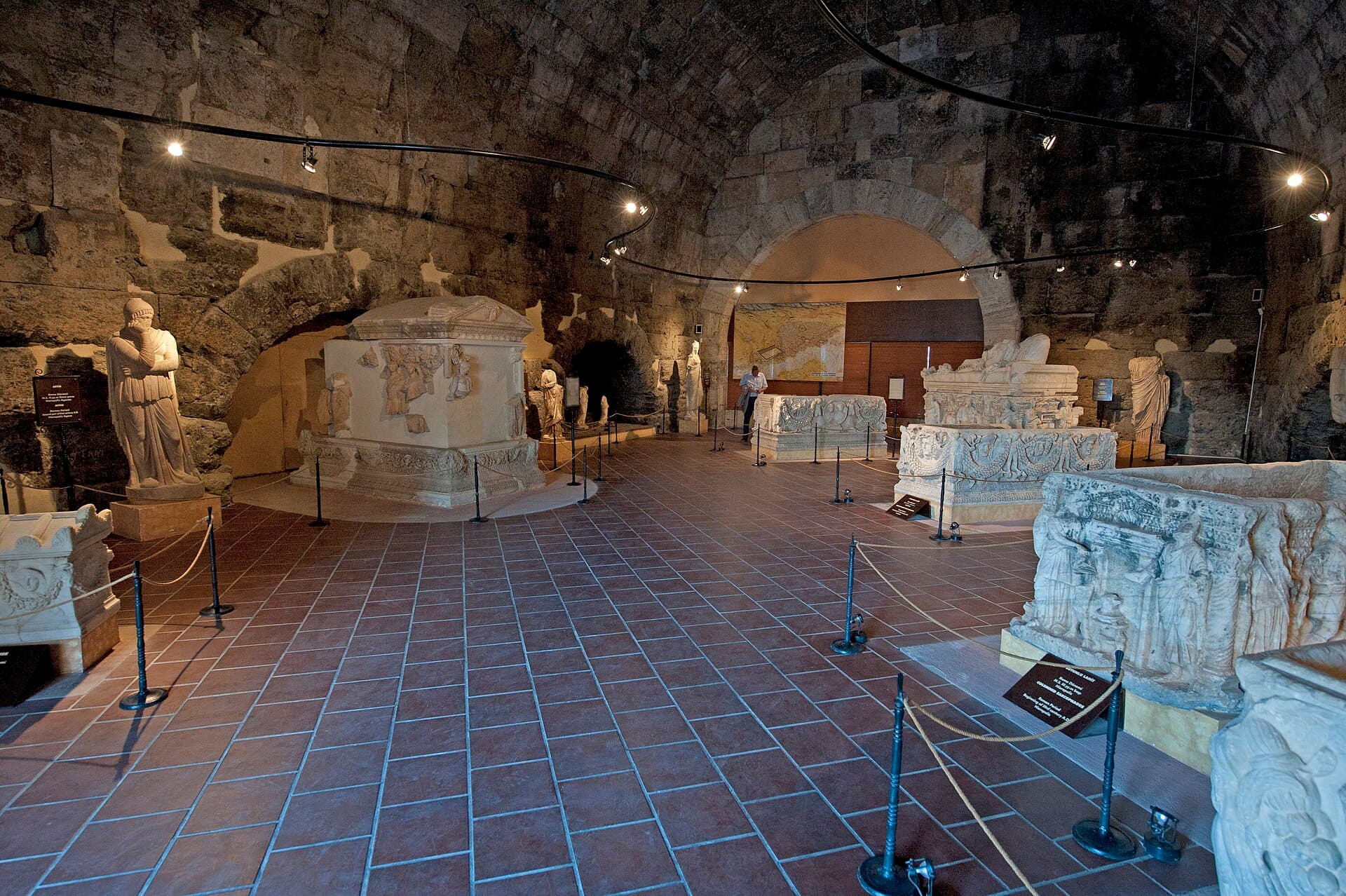 Garlanded Roman sarcophagi and marble statues stand under spotlights in a barrel-vaulted stone hall of the Hierapolis Archaeology Museum.