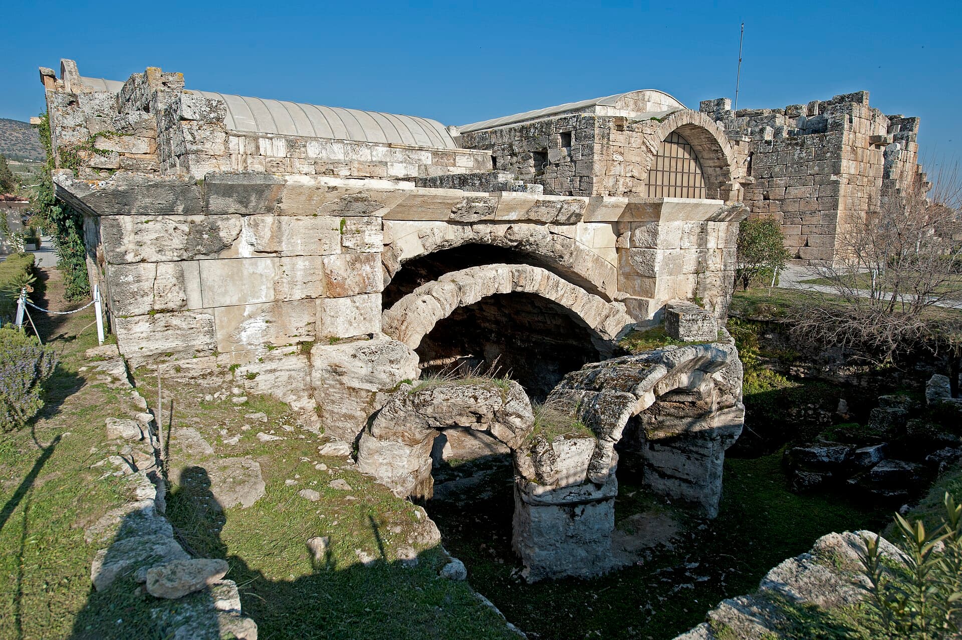 Massive stone walls and collapsed vault arches of the Roman bath complex that houses the Hierapolis Archaeology Museum, its restored hall with a great glazed arch standing behind.