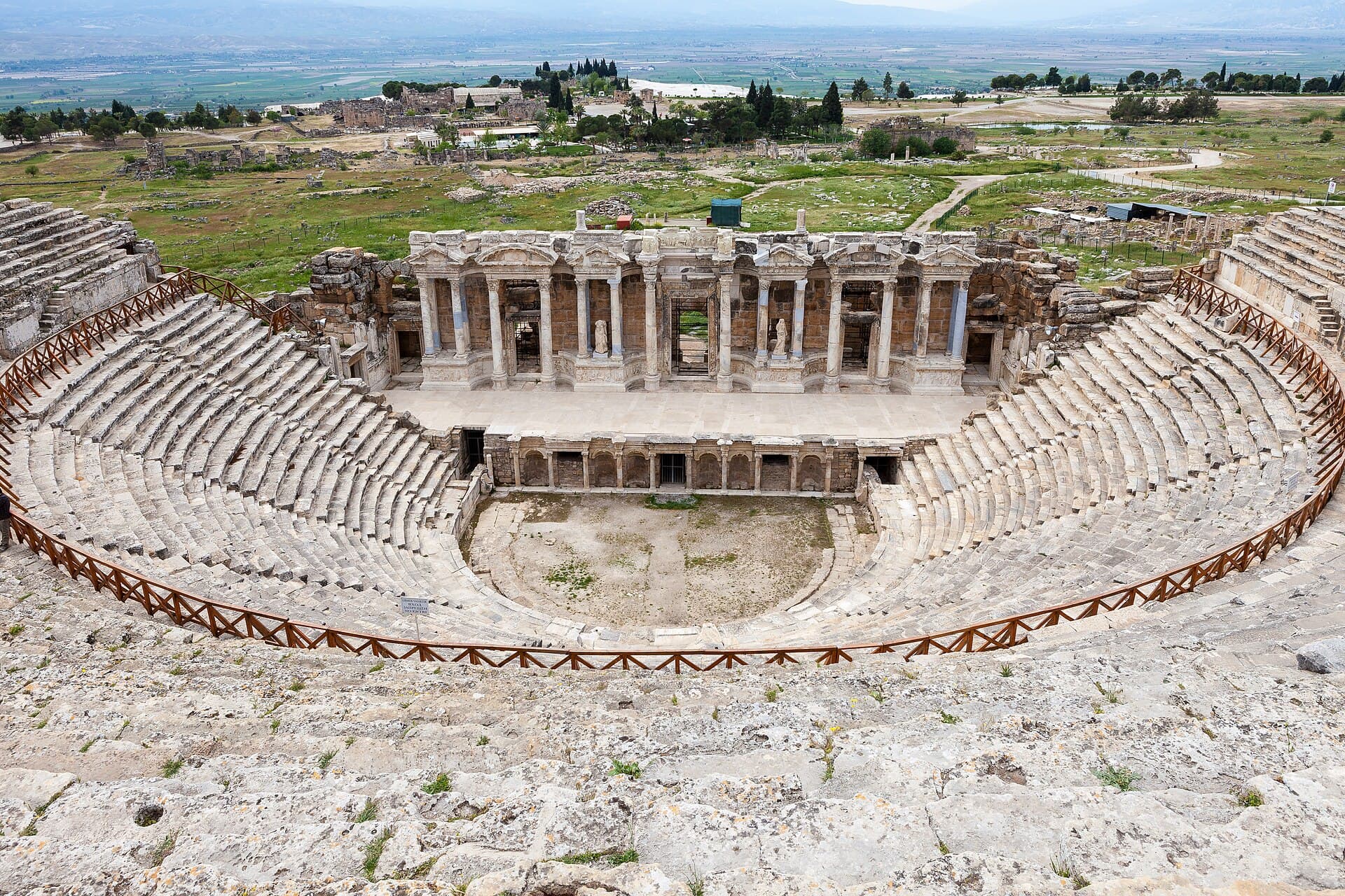 Seen from the upper rows, the semicircular stone cavea of the Hierapolis theatre curves around the orchestra toward the re-erected two-storey scaenae frons, whose marble columns and statue-filled niches stand against the green plain below.