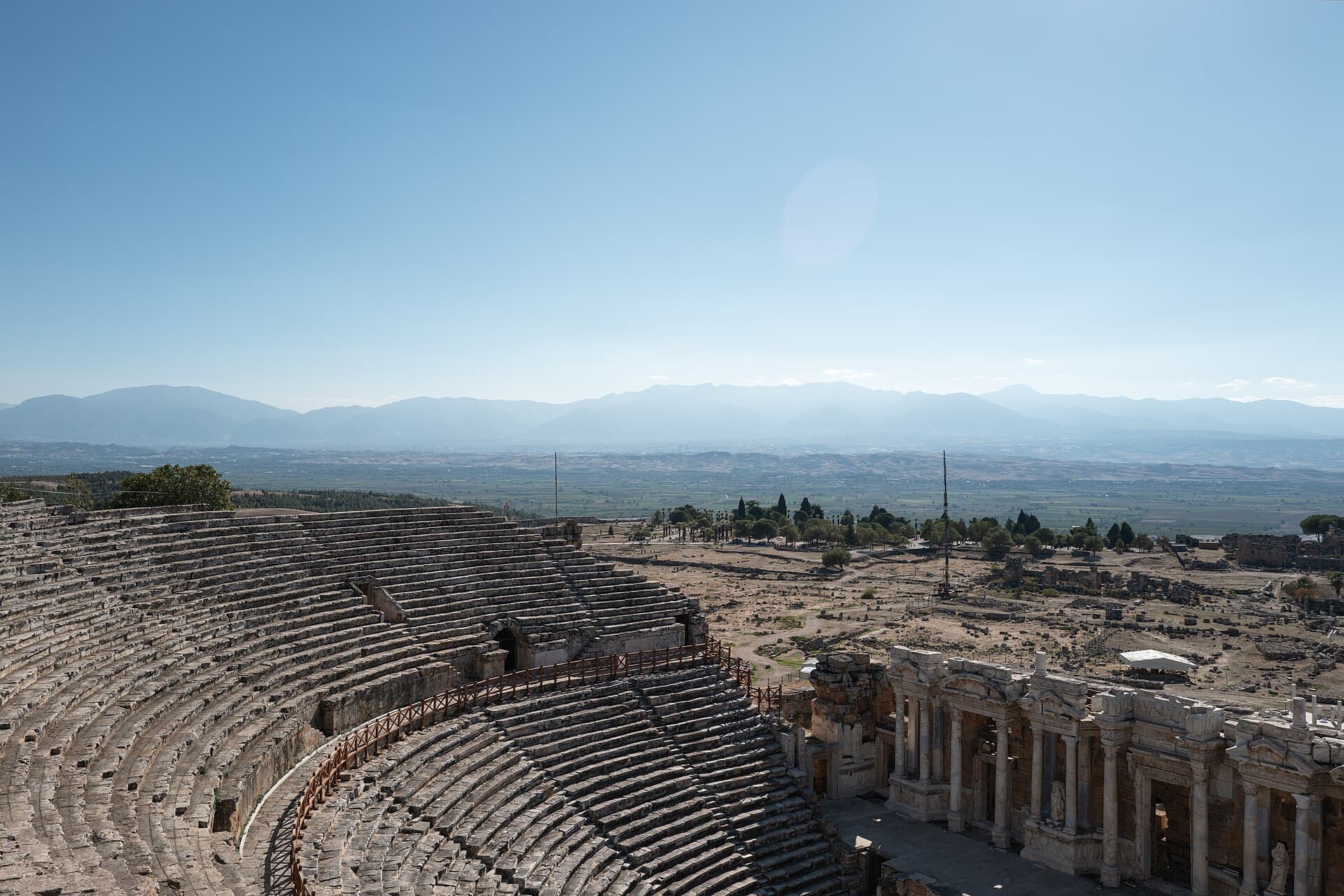From the topmost seating rows of the Hierapolis theatre, weathered stone tiers sweep down past the carved upper cornice of the stage building toward the scattered ruins of the ancient city and hazy mountains across the Lycus valley.