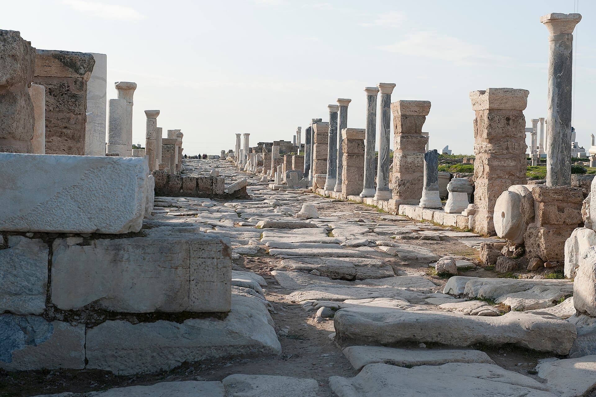 Worn stone paving of Syria Street at Laodicea stretches toward the horizon between rows of re-erected columns and masonry piers, with visitors visible far down the ancient colonnaded avenue.