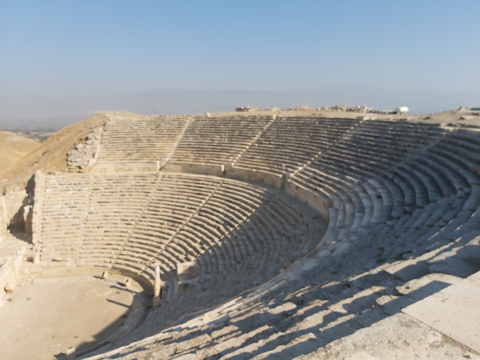 The restored semicircular seating tiers of the Western Theatre at Laodicea curve around its orchestra floor, seen from the upper rim with the hazy Lycus valley in the distance.