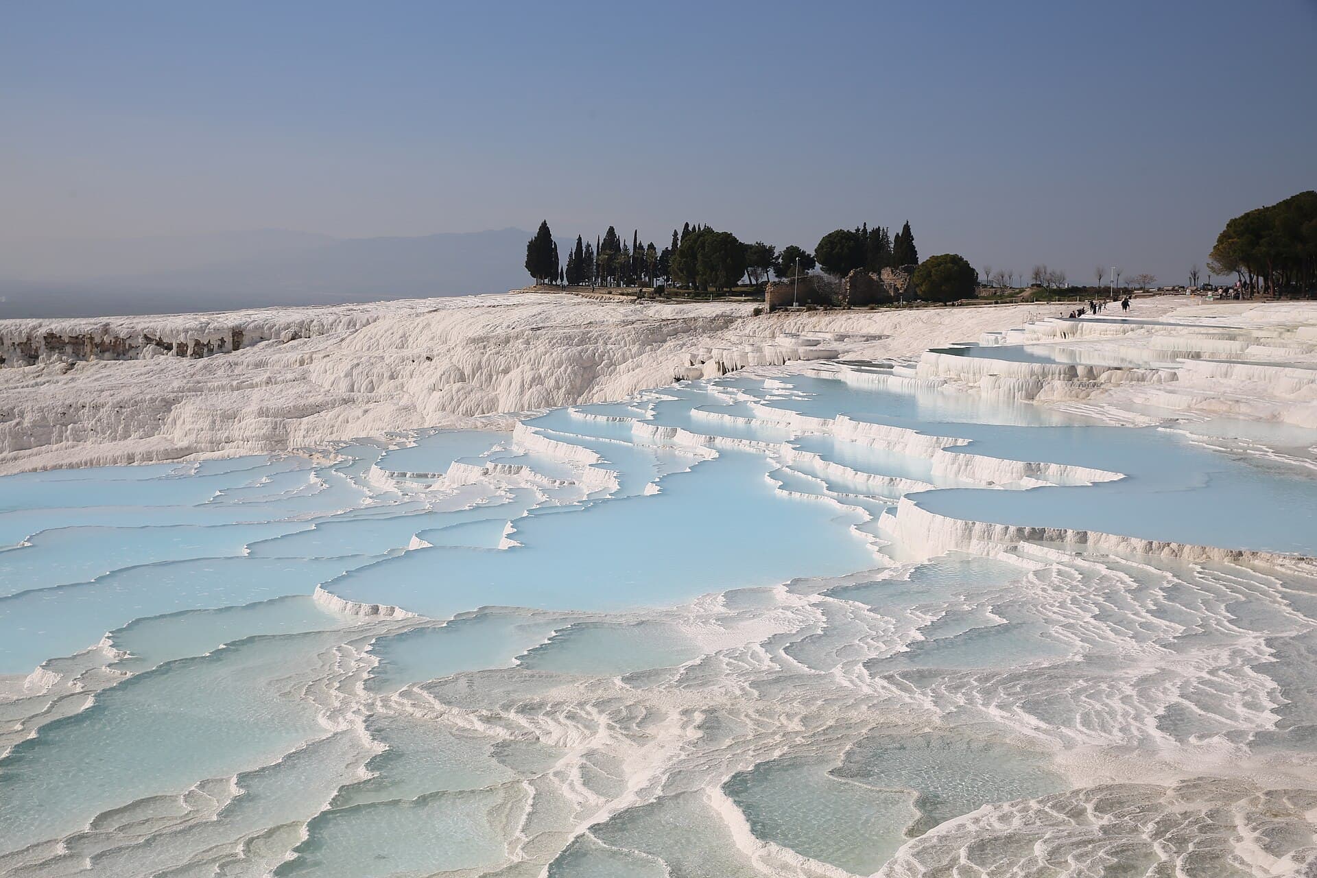 Terraced travertine pools at Pamukkale filled with shallow pale-blue water, their scalloped white rims stepping down toward the viewer, with trees and a few visitors on the ridge behind.
