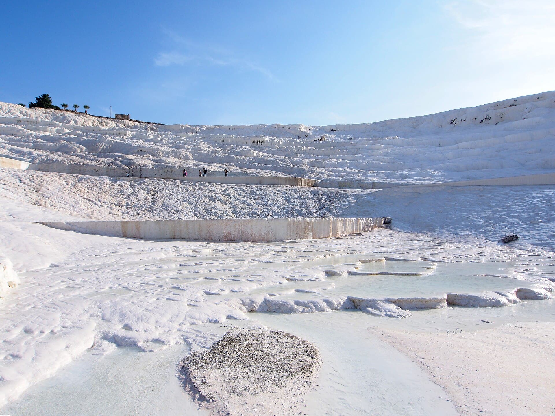 Looking up Pamukkale’s white travertine slope from its base, where a thin sheet of turquoise water covers only the lowest basins while the broad terraces above stand dry, with a few visitors walking far upslope.