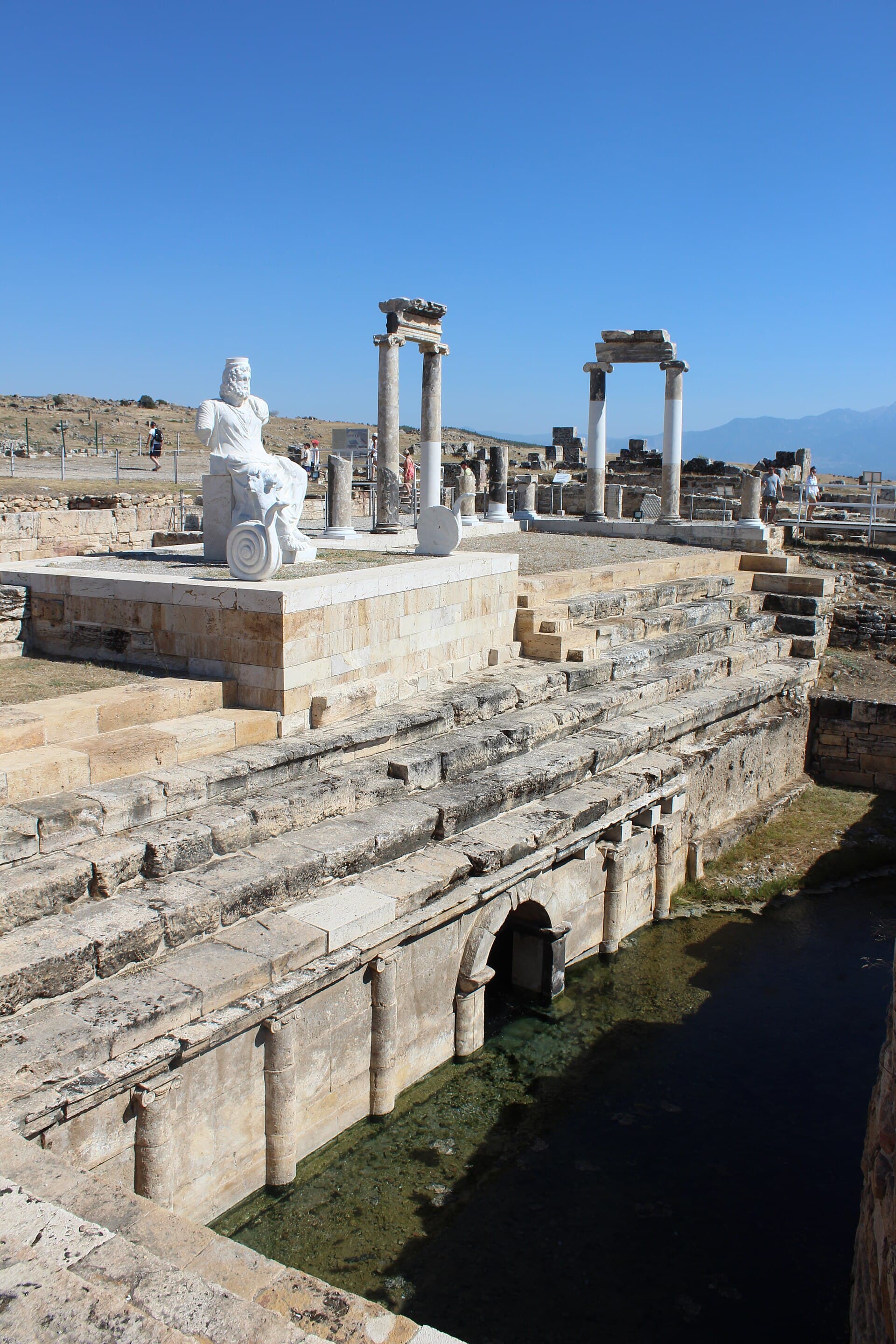 The excavated Ploutonion sanctuary at Hierapolis, with the white marble replica statue of Hades and Cerberus on its platform, re-erected columns, stepped spectator terraces, and the arched stone opening over the green thermal spring pool below.