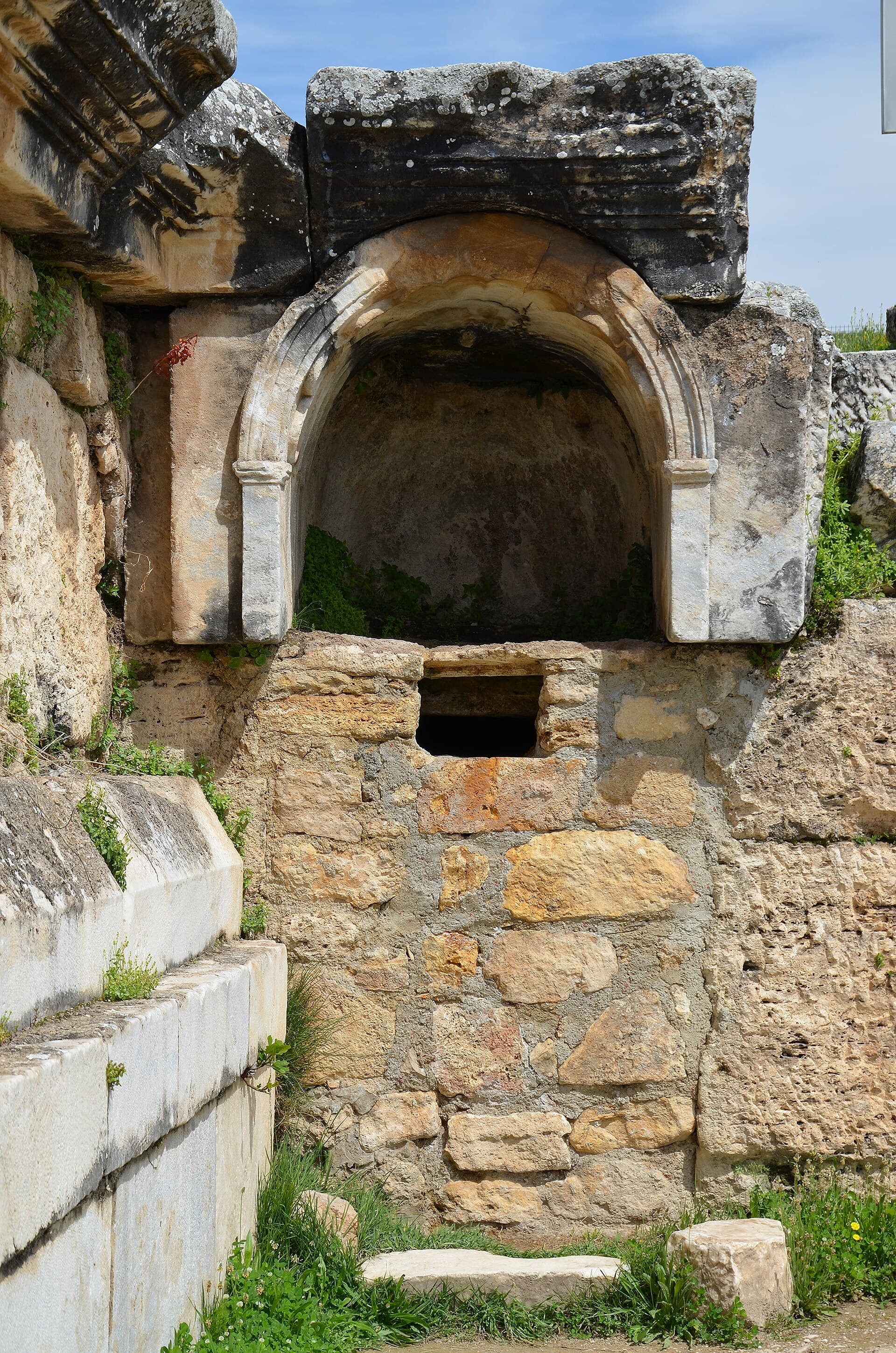 The marble arch of Pluto’s Gate at the Hierapolis Ploutonion, framing a shallow grotto niche above the small rectangular stone opening where toxic gases vent from the cave beneath.