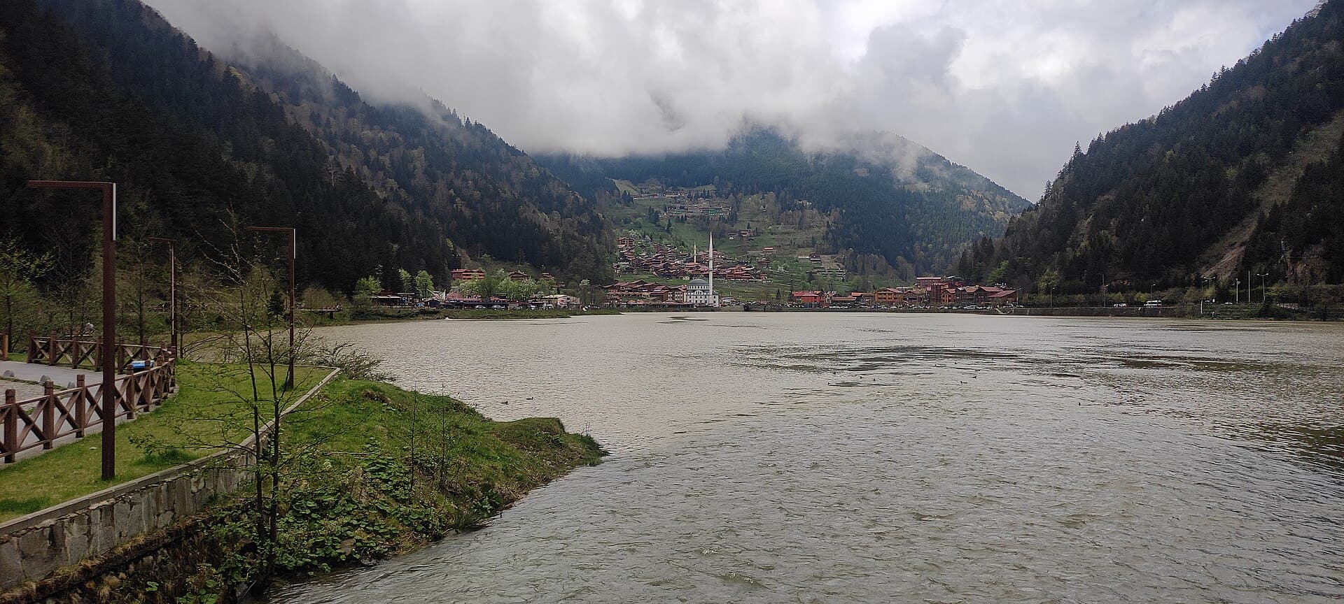 Lakeside boardwalk path along Uzungöl's shoreline with village and mountains beyond.